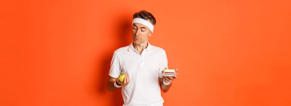 Portrait of handsome middle-aged man in good shape, wearing uniform for training, choosing between apple and cake, standing over orange background.