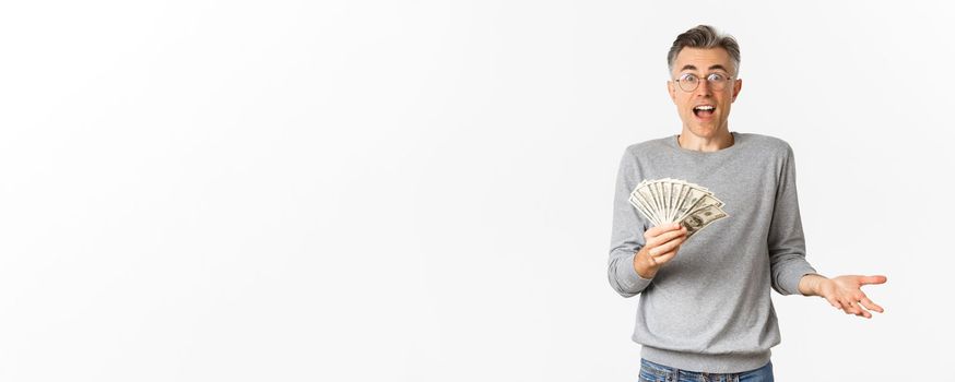 Portrait of surprised middle-aged man in glasses and gray sweater, holding money, winning lottery, standing over white background.