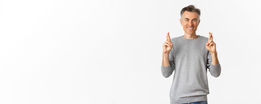 Image of hopeful middle-aged man making a wish, crossing fingers and smiling, anticipating good news, standing over white background.