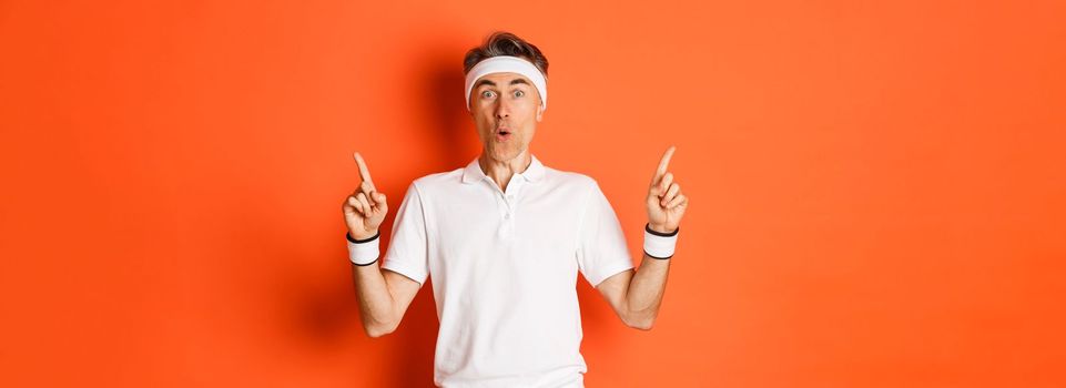 Portrait of handsome middle-aged guy in sports clothing, pointing fingers up, showing promo banner about gym or workout, standing over orange background.