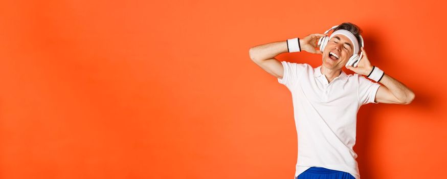 Portrait of carefree middle-aged male athlete, listening music in headphones during fitness training, standing over orange background.