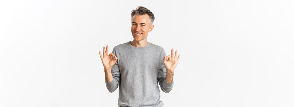 Portrait of handsome middle-aged man, smiling and looking confident while showing okay signs, guarantee something is good, standing over white background.
