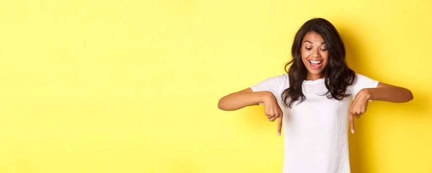 Portrait of excited, good-looking african american woman, pointing fingers down and looking with super happy face, standing over yellow background.