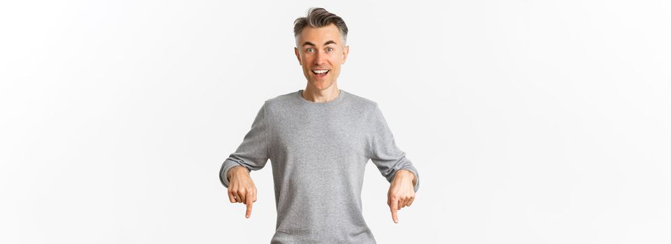 Image of surprised handsome middle-aged man pointing fingers down, smiling and looking curious, standing over white background in grey sweater.