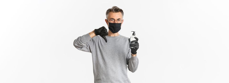 Concept of coronavirus, lifestyle and quarantine. Portrait of middle-aged man in medical mask and gloves do not recommend antiseptic, showing hand sanitizer and thumbs-down, white background.