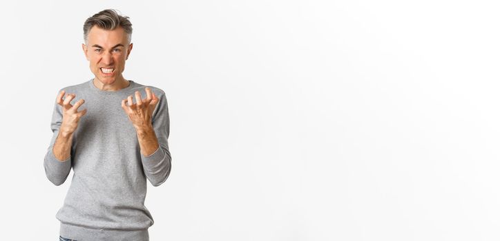 Image of angry middle-aged man cursing someone, looking mad and clenching fists with hatred, standing over white background.