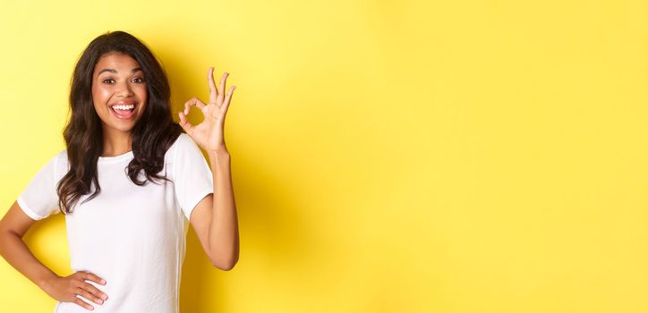 Portrait of happy good-looking african american girl, showing okay sign and smiling in approval, praise good work, standing over yellow background.