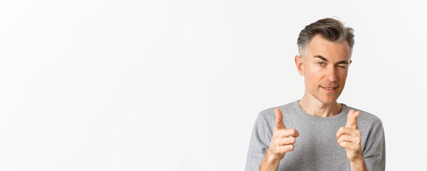 Close-up of handsome cheeky man with gray hair, winking and pointing fingers at camera, praising you, well done gesture, standing over white background.