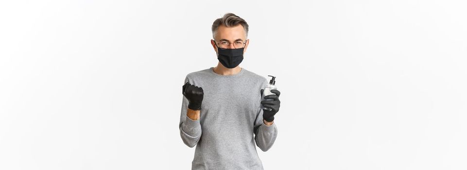 Concept of coronavirus, lifestyle and quarantine. Image of cheerful caucasian man in medical mask and gloves, showing hand sanitizer and making fist pump to rejoice, standing over white background.