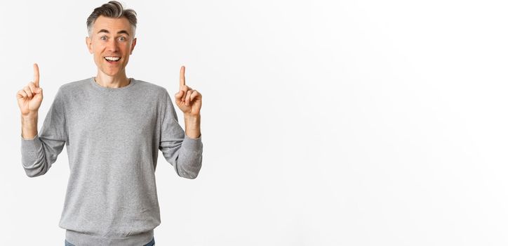 Image of amazed adult guy with grey short hairstyle, showing awesome offer, pointing fingers up at copy space and smiling happy, standing over white background.