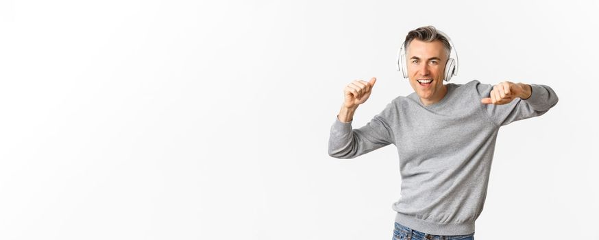 Portrait of carefree, modern middle-aged man, listening music in headphones and dancing, standing over white background.