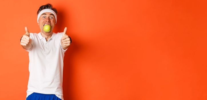 Image of funny middle-aged guy in fitness clothing, holding apple in mouth, showing thumbs-up, standing over orange background.