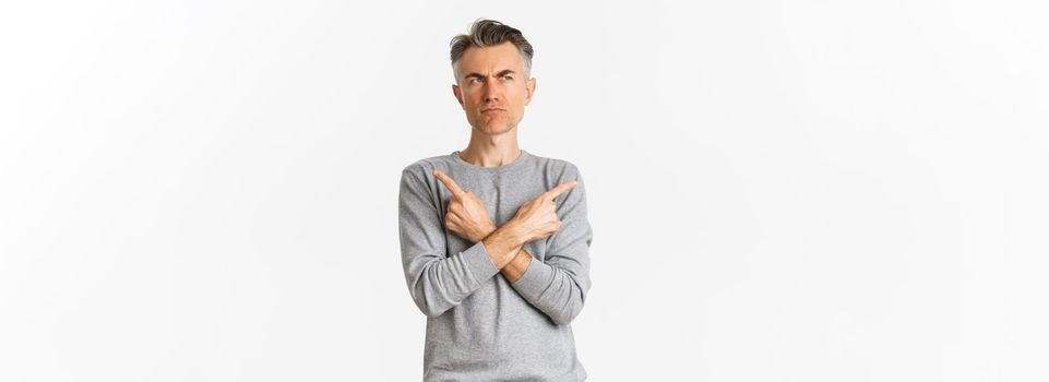 Image of indecisive middle-aged man making choice, pointing fingers sideways and frowning doubtful, standing unsure over white background.