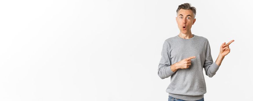 Amazed handsome guy in grey sweater, pointing fingers right and looking curious, askign question about product, standing over white background.