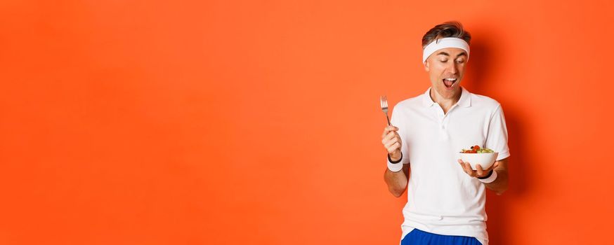 Concept of sport, fitness and lifestyle. Portrait of joyful middle-aged guy in workout uniform, holding fork and salad, eating healthy food, standing over orange background.