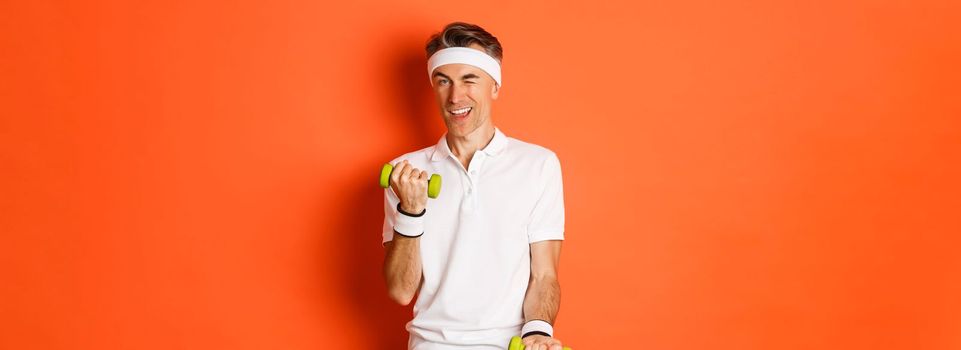 Portrait of handsome cheeky, middle-aged male athlete doing sports, exercise with dumbbells and winking at camera, standing over orange background.