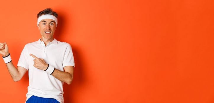 Portrait of amazed middle-aged male athlete, pointing fingers left and smiling, showing logo about gym or workout, standing over orange background.