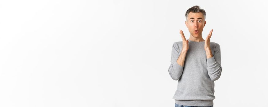 Image of middle-aged man with gray short hair, looking amazed and fascinated, saying wow, reacting to advertisement, standing over white background.