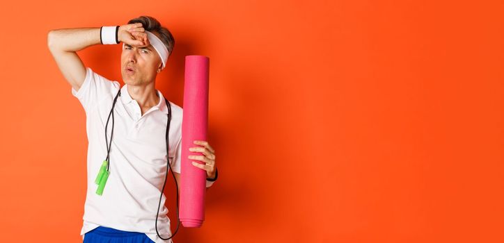 Concept of workout, gym and lifestyle. Image of handsome middle-aged man, tired after fitness exercises, holding skipping rope and yoga mat, wiping sweat off forehead, orange background.