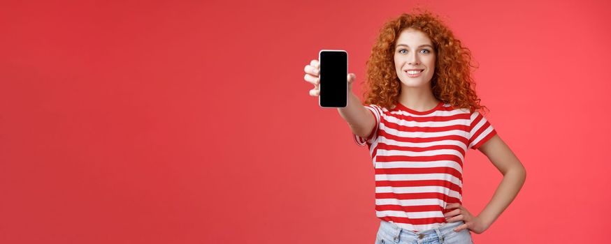 Confident good-looking redhead curly woman present cool app hold smartphone extended hand show phone screen smiling assertive recommending subscribe her blog social-media. Copy space