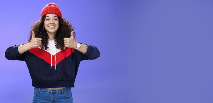 Girl totally agrees recommends cool place showing thumbs-up and smiling excited with happy grin standing upbeat and delighted against blue background in warm beanie and sweatshirt. Body language and emotions concept