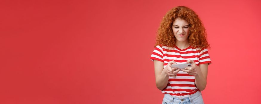 Daring enthusiastic addicted geeky good-looking redhead curly girl hate losing battles smartphone game wrinkle nose grimacing upset eager win tap fast telephone screen look display, red background.