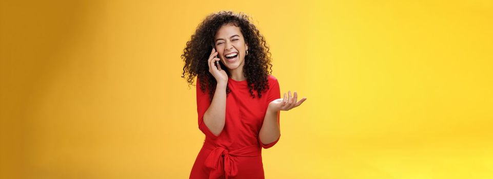 Portrait of joyful charming european female in red dress with curly hair laughing out loud as gossiping with friend via smartphone close eyes as giggling gesturing, hearing joke through mobile phone.