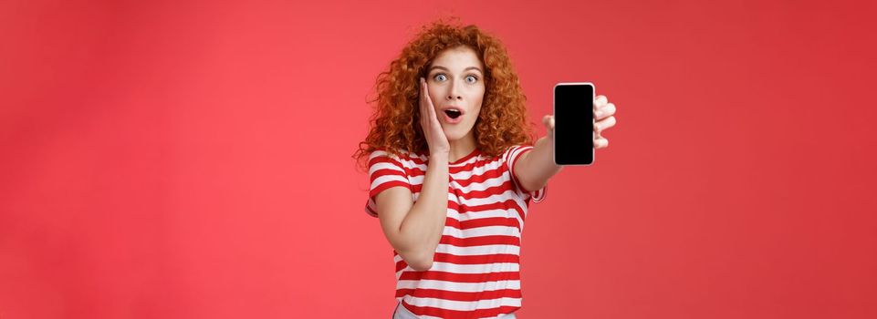 Lifestyle. Amused impressed redhead good-looking curly-haired excited girl share awesome social media page show smartphone screen amazed touch cheek thrilled affected awesome app red background.