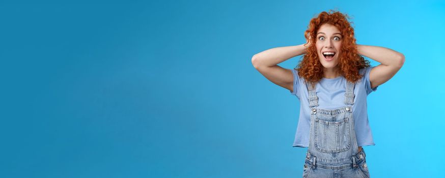 Lifestyle. Excited amused joyful redhead curly girl having amazing perfect news touch hair astonished stare camera thrilled cheerful overjoy smiling broadly toothy carefree grin denim overalls blue background.