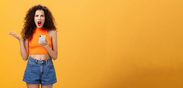 Portrait of displeased pissed and irritated caucasian female. with curly hairstyle in red lipstick and orange cropped top holding smartphone and gesturing with palm annoyed complaining on dumb message.