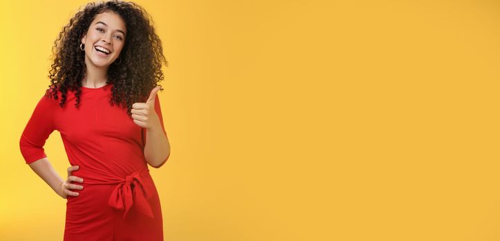 Portrait of happy optimistic young curly-haired woman in red dress laughing joyfully, showing thumbs up in approval and like gesture, delighted with awesome idea, accepting plan over yellow wall.