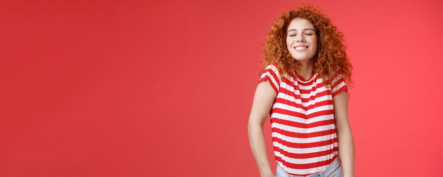 Cheerful relaxed carefree happy attractive redhead curly woman close eyes enjoy sunlight warm summer holiday weekends smiling broadly spend vacation sea trip travel joyfully, red background.