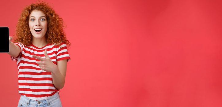 Take look what awesome smartphone. Impressed excited good-looking female love digital innovations redhead girl curly hairstyle showing phone screen pointing display promote cool app.