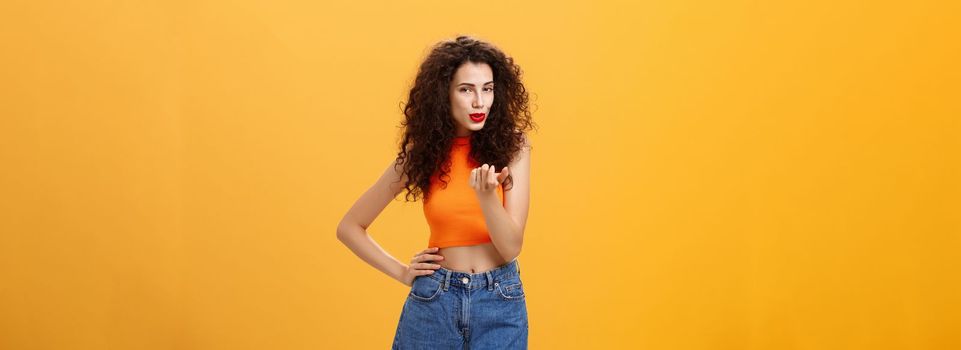 Woman seducing hot guy. Sensual and sexy daring female with curly hairstyle in stylish modern cropped top and shorts showing come here gesture inviting move closer flirting over orange background.