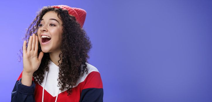 Friend shouting through street to turn attention. Portrait of carefree happy cute woman in beanie and sweatshirt holding palm near opened mouth looking with smile left as calling mate with loud yell.