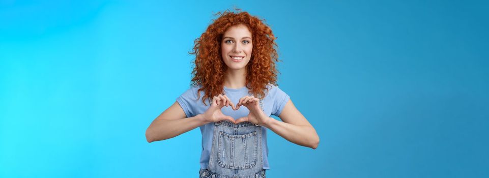 Tenderness, love, romance concept. Lovely feminine cute redhead curly woman show heart gesture chest express sympathy passionate feelings smiling broadly cherish feelings standing blue background.