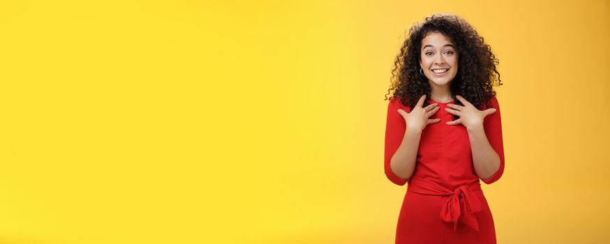 Lifestyle. Pleased and thankful tender curly-haired kind girl in red dress pressing hands to breast being surprised with unexpected tender heartwarming gift thanking smiling delighted over yellow background