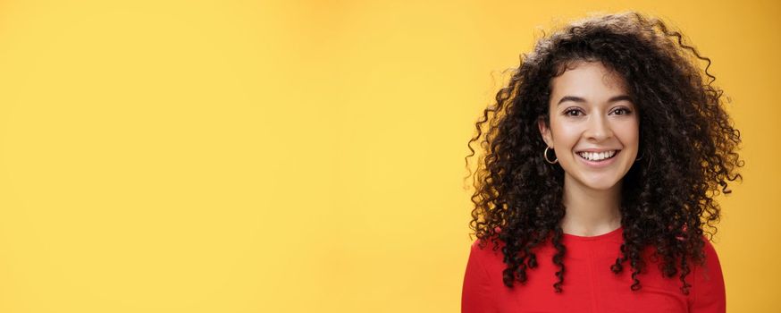 Close-up shot of pretty caucasian girl with curly hair in red dress and earrings smiling joyfully with pleased hopeful expression gazing at camera carefree, posing over yellow background.