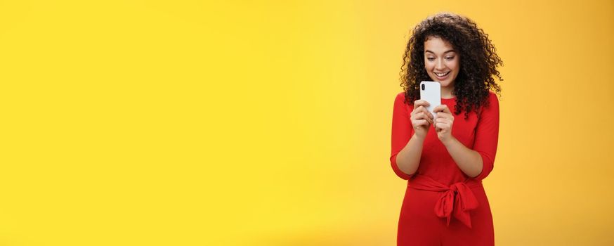 Wow new mobile phone amazing. Impressed and astonished good-looking curly-haired female in red dress holding smartphone looking at screen amused as playing cool app or game over yellow wall.