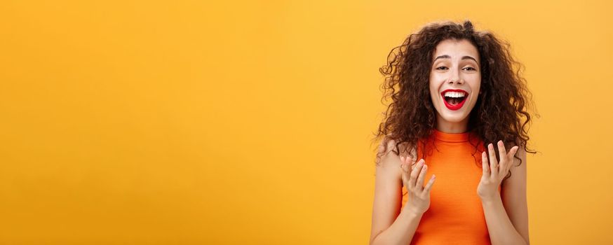 Surprised thrilled and joyful charming european female. with curly hairstyle raising palms in grateful and delighted gesture smiling broadly hearing unbelievable perfect news over orange wall. Copy space