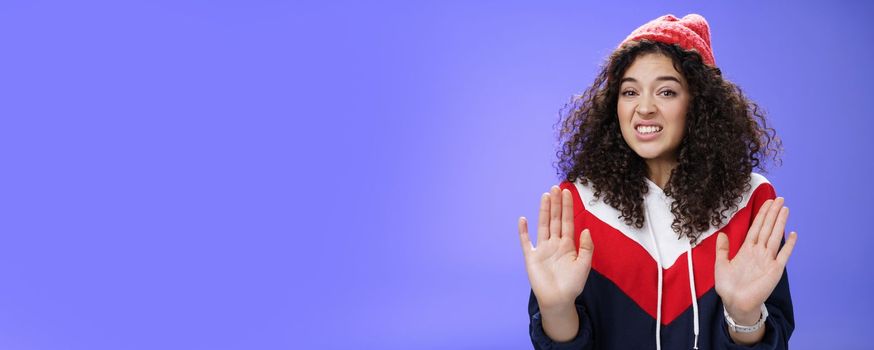 Lifestyle. Displeased european woman with curly hair raising palms near chest and waving in refusal and rejection gesture grimacing unsatisfied and unwilling to participate in suspicious action over blue wall.