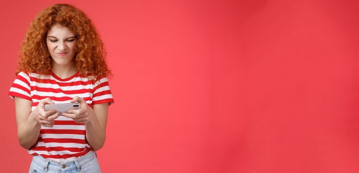 Daring enthusiastic addicted geeky good-looking redhead curly girl hate losing battles smartphone game wrinkle nose grimacing upset eager win tap fast telephone screen look display, red background.