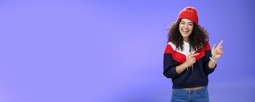 Charismatic playful young curly-haired european female winking joyfully at camera as pointing at upper right corner wearing hat and pullover as posing delighted over blue background, feeling warm.