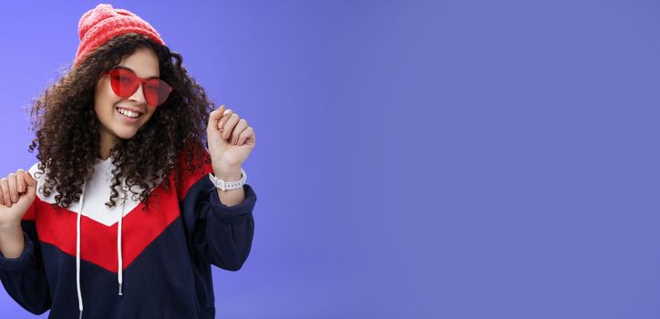 Lifestyle. Stylish and feminine girlfriend dancing having fun feeling upbeat as liking weather feeling warm and cozy in stylish red beanie and sunglasses dancing with raised hands and broad smile over blue wall.