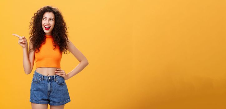 Portrait of delighted enthusiastic stylish young woman. with curly hairstyle in red lipstick and cropped top holding hand on waist pointing and looking left amused and happy over orange background.
