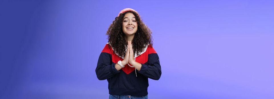 Lifestyle. Charming excited and friendly-looking cute female friend with curly hairstyle in outdoor hat and sweatshirt holding hands in pray and smiling with angel expression as hoping friend help over blue wall.