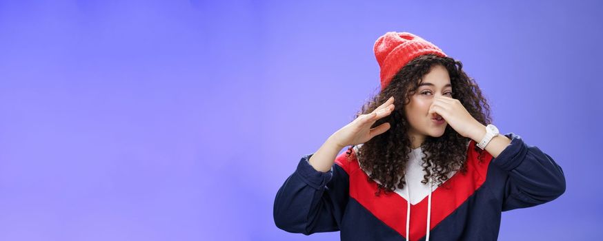 Gosh what stink. Portrait of cute caucasian girl with curly hair in cute trendy hat close nose with fingers and waving hand as smelling awful reek or smell standing disgusted against blue background.