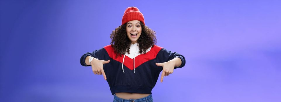 Hurry up and chek it out. Portrait of charismatic joyful and excited young cute curly-haired girl in beanie and stylish outfit pointing down, smiling broadly inviting join fun over blue background.