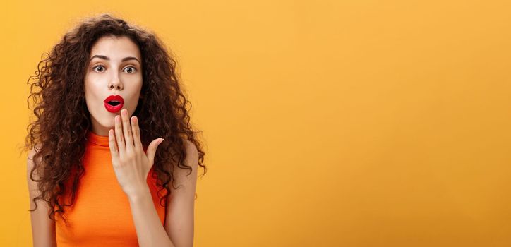 Waist-up shot of amazed and excited caucasian woman. with curly hair in red lipstic opening mouth from surprise covering it with palm listening shocking rumors reacting astonished to amusing news.