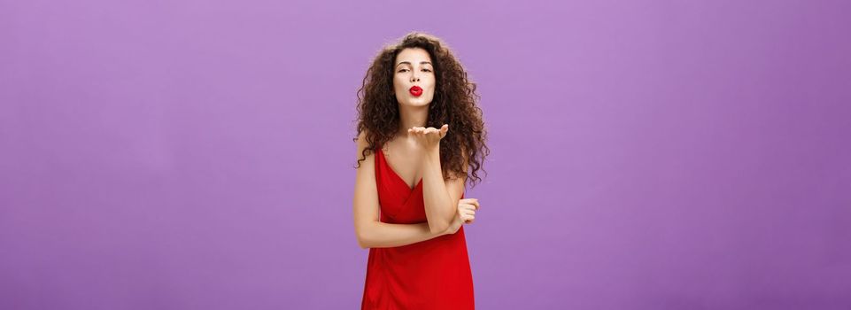 Woman sending passionate kiss being hopeless romantic. standing in elegant red dress with curly hairstyle and makeup blowing mwah with smile and folded lips holding palm near mouth over purple wall.